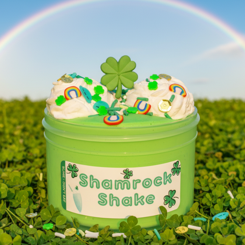A light green container labeled 'Shamrock Shake' filled with fluffy slime decorated with shamrock and rainbow sprinkles, a peppermint candy charm, and two little clay 'whip cream' pieces.