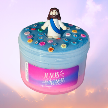 A container of 'Jesus is Risen - Butter Slime' with a colorful mix of pink, blue, and purple slime, topped with a charm of Jesus and flower and cloud sprinkles against a sunset background.
